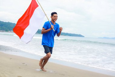 Genç bir adam Endonezya Bağımsızlık Günü 'nü kutluyor. Ulusal bayrağıyla sahilde koşuyor.