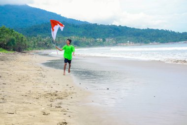Genç adam, Endonezya bayrağı rüzgarda dalgalanırken sahilde neşeyle koşar.