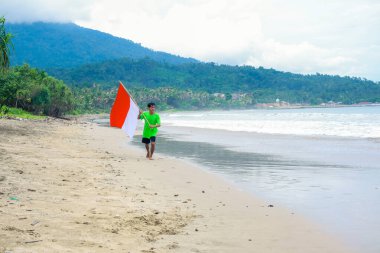 Genç bir adam elinde Endonezya bayrağıyla plaj boyunca koşup bağımsızlık gününü kutluyor.