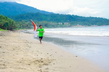 Endonezya bayrağıyla plajda koşan genç adam, bağımsızlık gününü kutluyor.