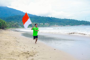 Genç bir adam sahilde neşeyle koşuyor, Endonezya bayrağını sallıyor.
