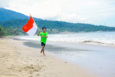 Genç bir adam Endonezya 'nın Bağımsızlık Günü' nü kutluyor. Plajda koşuyor ve elinde bayrak sallıyor.