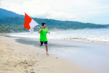 Genç bir adam Endonezya bayrağıyla sahilde neşeyle koşuyor, ulusal gururu simgeliyor.
