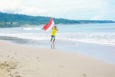 Genç adam sevinçle sahilde koşuyor, Endonezya Bağımsızlık Günü 'nü coşkulu bir bayrağla kutluyor.