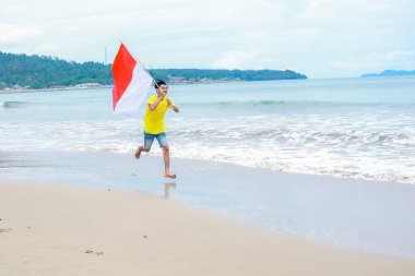 Genç bir adam neşe içinde Endonezya bayrağıyla tropik bir sahilde koşuyor..
