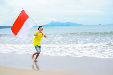 Genç adam sevinçle sahilde koşuyor, Endonezya bayrağını gururla sallıyor.