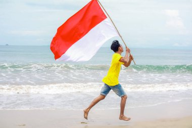 Bağımsızlık Günü 'nde kumlu bir plaj boyunca Endonezya bayrağıyla koşan genç bir adam.
