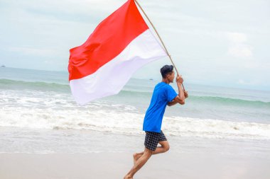 Adam sahilde koşuyor, gururla Endonezya bayrağını sallıyor, bağımsızlık gününü kutluyor.