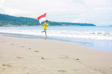 Endonezya bayrağını gururla taşıyan bir adam kumlu bir sahilde neşeyle koşar..