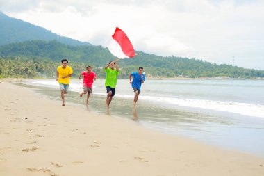 Grup, Endonezya Bağımsızlık Günü 'nü ulusal bayrağımızla kutlayarak, tropikal bir sahilde neşeyle koşuyor.