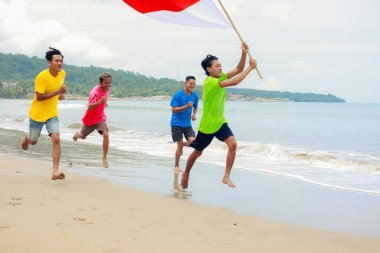 Bağımsızlık Günü 'nü kutlayan bir grup, Endonezya bayrağıyla tropik bir plaj boyunca neşeyle koşuyor.