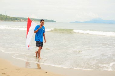 Neşeli genç adam ulusal günü kutluyor, Endonezya bayrağıyla sakin bir plaj boyunca yürüyor..