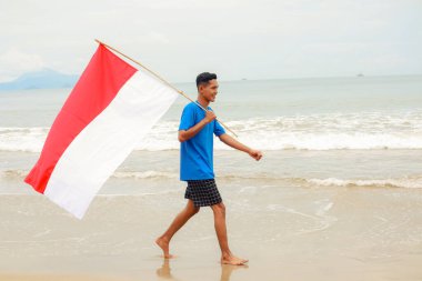 Bağımsızlık gününü kutlayın. Endonezya bayrağı taşıyan genç bir adam sahilde yürüyor.