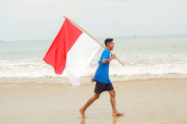 Genç adam sahilde yürüyor, Bağımsızlık Günü 'nü kutlayan Endonezya bayrağını neşeyle taşıyor.