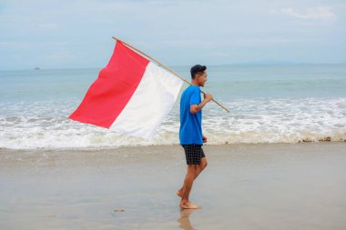 Genç Endonezyalı bir adam güzel bir sahilde bayrağıyla bağımsızlık gününü kutluyor.
