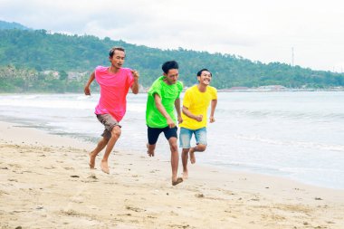 Renkli gömlekli üç genç kumsalda neşeyle koşuyor.
