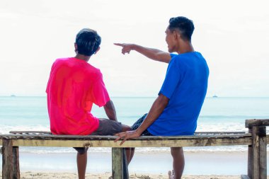 İki Renkli Gömlekli Adam Sahilde dinleniyor Denizi işaret ediyor