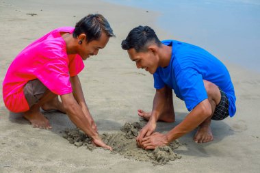 Güneşli bir sahilde kumdan kale inşa eden iki adam.