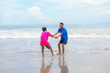 İki genç adam, Güneşli bir plajda el ele tutuşup koşuyor.