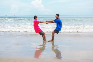 Güneşli bir sahilde el ele tutuşan ve gülümseyen iki adam.
