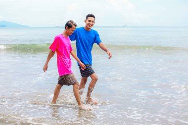 Renkli gömlekli iki adam güneşli bir sahil boyunca neşeyle yürüyor.