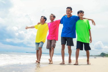 Güneşli bir plajda yürüyen dört renkli gömlekli genç adam.