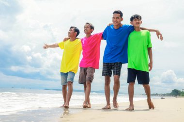 Renkli gömlekli dört genç güneşli bir sahilde neşeyle yürüyor.