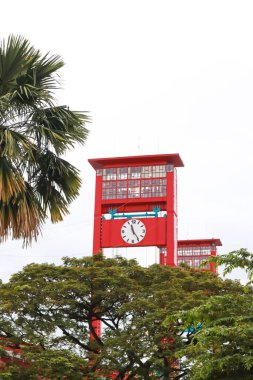 Canlı bir kırmızı saat kulesi, şehir manzarasında yemyeşil ile çevrili, uzun duruyor.