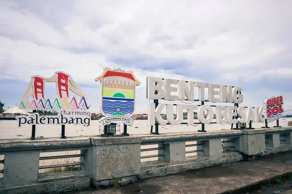 Bulutlu bir gökyüzünün altında ikonik simgelerle Palembang 'ın nehir manzarasını gösteren göz seviyesinde bir görüntü..