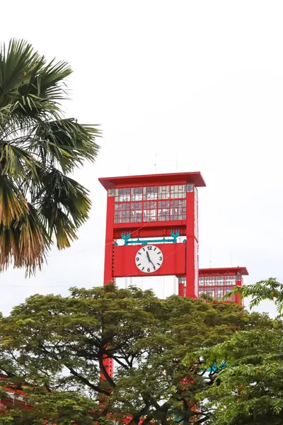 Canlı bir kırmızı saat kulesi, şehir manzarasında yemyeşil ile çevrili, uzun duruyor.