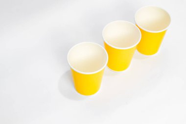Beyaz arka planda çaprazlama dizilmiş üç parlak sarı kağıt bardak yumuşak gölgeler oluşturuyor ve minimalizmi gözler önüne seriyor.