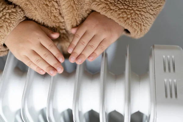 Child wearing warm clothing warms hands on heaters on a cold winter day at home.