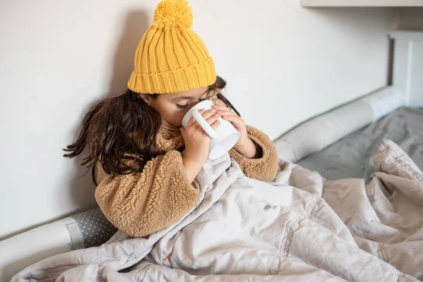 Stock image Beautiful little girl warms in blanket in cold winter time. Child in warm clothes sitting in a bed drinking hot tea at home. Cold home. No heating.