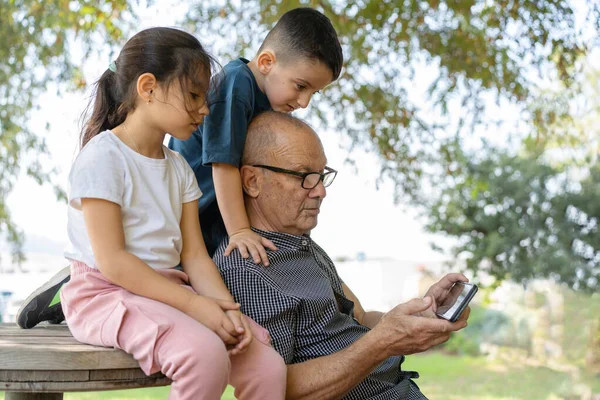 Happy family, joyful little children hugging grandfather and using smart phone while sitting together outdoors on nature background.