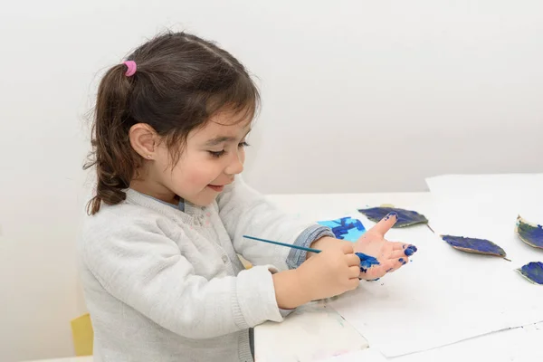 Little girl painting leaves blue color, crafts and art therapy. Child paints her own hand with blue paint and paintbrush getting messy and having fun.