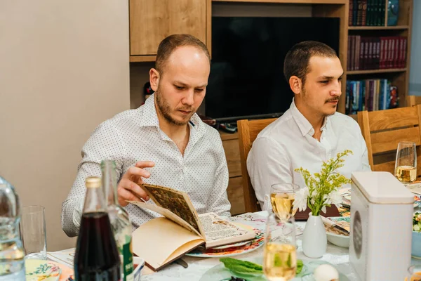 Passover Seder.Two Men Celebrate Passover with Traditional Foods, and Reading Haggadah. Jewish Family At The Table Celebrating Pesach. Young Bearded Man Reads The Passover Haggadah.