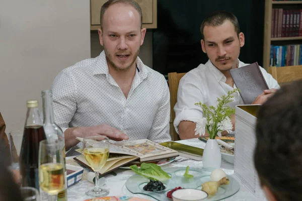 Passover Seder.Two Men Celebrate Passover with Traditional Foods,Reading Haggadah.Jewish Family Celebrating Pesach.Young Bearded Man Reads The Passover Haggadah.Traditional seder plate on table.