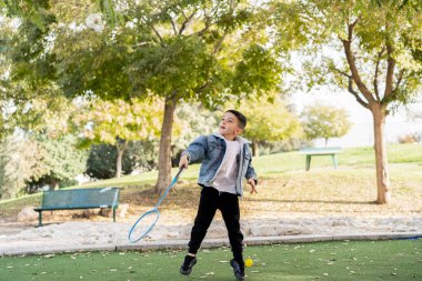 Çocuk Sıçrıyor ve Shuttlecock 'a raketle vuruyor. Aktif Çocuk, Sunny City Park 'ta Badminton oynuyor, Yeşil Çim Sahası' nda raket ve Shuttlecock ile eğleniyor, Doğa Arkaplanı.