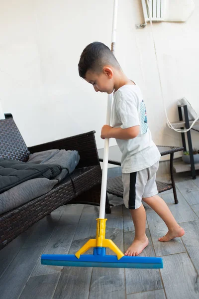 Funny young child cleaning floor at home. Boy washing floor with floor squeegee in balcony of modern apartment. Male household helping tidy house.