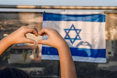 Childs Tender Hands Create a Heart Shape, Sunlight Casting Form Heart Shadow on the Israeli National Flag Hanging On the Balcony. clipart