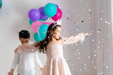 Pembe elbiseli bir kız, doğum günü partisinde etrafı konfeti ve renkli balonlarla çevrili kardeşiyle neşeyle dans eder..