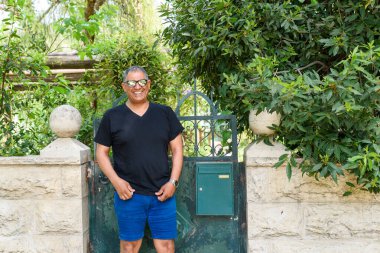 Günlük siyah tişörtlü ve mavi şortlu neşeli son sınıf öğrencisi yeşil kapının yanında duruyor. Bahçesi sakin, yemyeşil. Seyahat, turizm ve açık hava eğlencesi temaları için mükemmel.