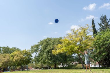 Parkta güneşli bir günün tadını çıkaran küçük bir çocuk, parlak gökyüzüne mavi bir frizbi fırlatırken yemyeşil ağaçlar ve açık mavi bir zemin ile çevrili..