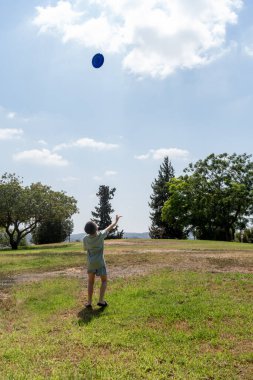 Küçük çocuk yeşil bir parkta mavi bir frizbi fırlatıyor. Parlak gökyüzü ve ılık güneş, arka planda gölgeler bırakan ağaçlar gibi neşeli bir atmosfer yaratıyor. Dışarıda eğlenmek için mükemmel bir gün..