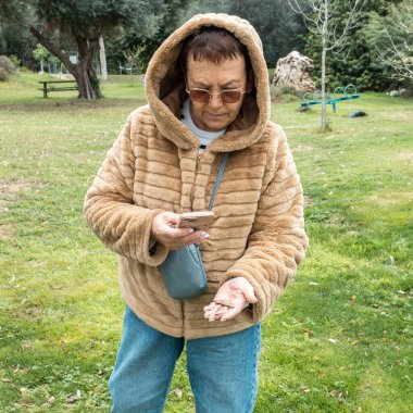 Son sınıf öğrencisi bir kadın, muhtemelen yapay zeka uygulaması olan akıllı bir telefon kullanıyor, parkta yürürken yerde bulunan akçaağaç tohumlarını incelemek için. Teknoloji şu anda doğayla iç içe..