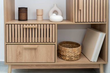 Şık bir ahşap depolama ünitesinde süslemeli vazolar ve örülmüş bir sepet bulunan çekmeceler ve raflar yer alıyor. Parlak odada nötr tonlu minimalist bir estetik sergileniyor..