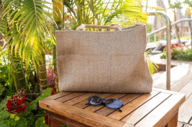 Havalı bir sahil çantası ve siyah güneş gözlükleri güneşli bir tatil beldesinde, tropik bitkilerle çevrili ahşap bir masanın üzerinde dinleniyor, rahat bir tatil atmosferini çağrıştırıyor..