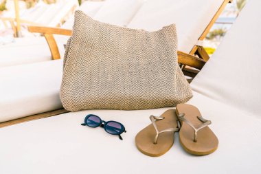 Plaj çantası, güneş gözlüğü ve parmak arası terlikler, sandalyeler arasında bir damla su olan temiz beyaz bir şezlongda dinleniyor. Deniz ya da havuz kenarındaki bir tatil köyünde şık ve sakin bir yaz tatili..