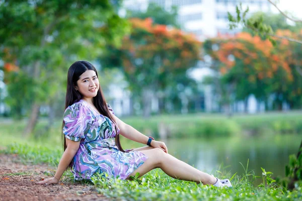 幸せなリラックスした肖像アジアの女性屋外市内公園の貯水池の横にある緑の芝生の芝生の上に座っている間 スマートウォッチを身に着けている紫色のドレス — ストック写真