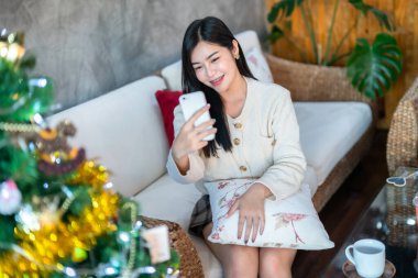 Güzel, güzel, pozitif gülümseyen Asyalı bir kadın akıllı telefon görüntülerini kullanıyor ya da Noel ve yeni yıl tatillerinde evde oturma odasındaki dekorasyonda selfie çekiyor..